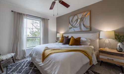 Bright bedroom with a large upholstered bed, wooden nightstands, bedside lamps, a ceiling fan, neutral décor, and a window providing natural light.